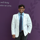 Suhas Babu in his white coat in front of the Kansas City University sign that reads: improving the well-being of the communities we serve